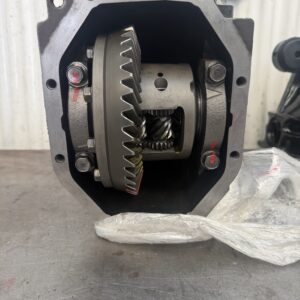 A close-up view of an open metal gearbox or differential, showing gears and mechanical components inside, resting on a workbench with a plastic sheet and a wall-mounted paper in the background.