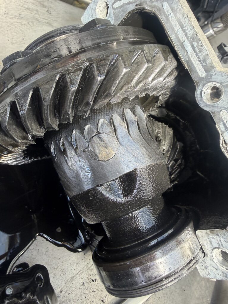 Close-up of a damaged automotive differential gear with visible metal wear and chipped teeth, surrounded by leaked black oil inside the opened metal housing.