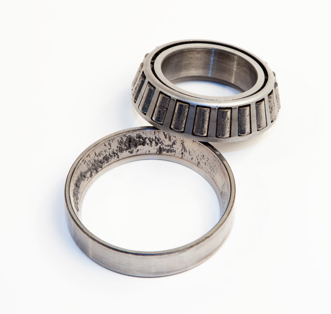 A close-up of a worn tapered roller bearing and its matching race, both showing signs of pitting and damage, placed on a white background.