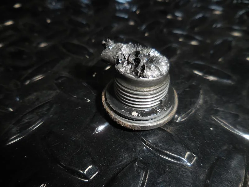 A close-up of a damaged, burnt metal bolt with melted and charred material on top, resting on a textured black surface.