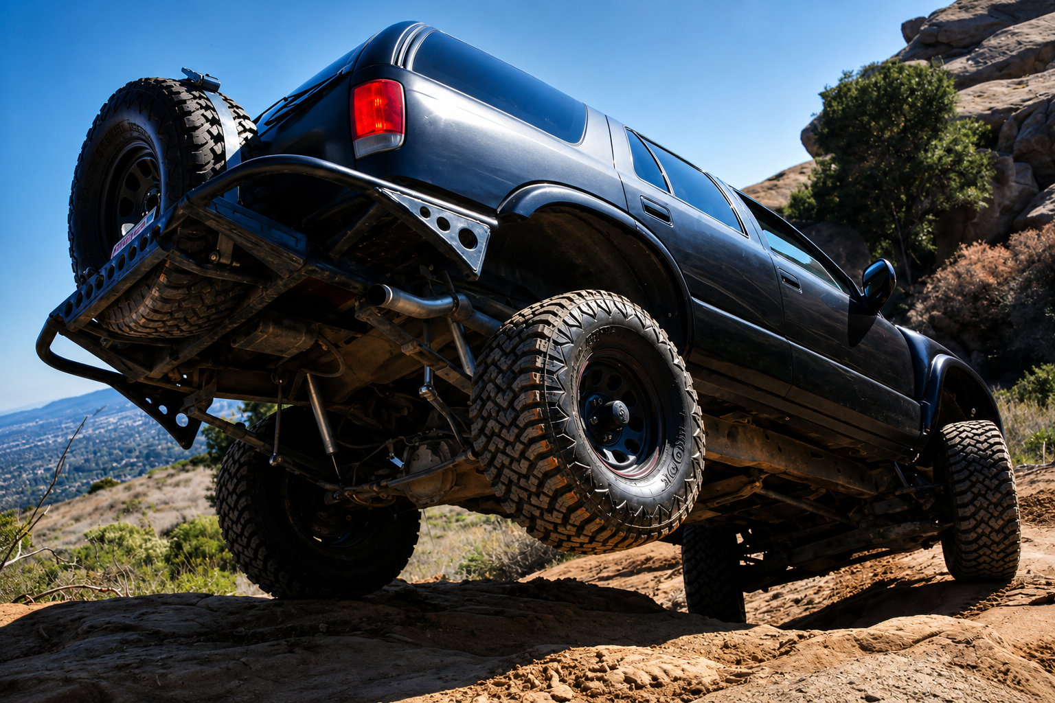 A black off-road 4x4 with large, rugged tyres climbs over rocky terrain on a sunny day, displaying its suspension system and elevated ground clearance. Bushes and hills are visible in the background.