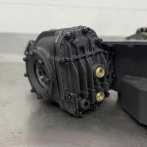 A close-up of a black mechanical gearbox or differential with multiple bolts and two brass-coloured fittings, resting on a metal surface with a white wall in the background.