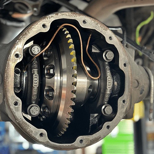 Close-up of an open differential gear assembly in a vehicle, showing shiny metal gears, bolts, and a thin copper tube inside the housing. Background is blurred with hints of machinery and green colour.