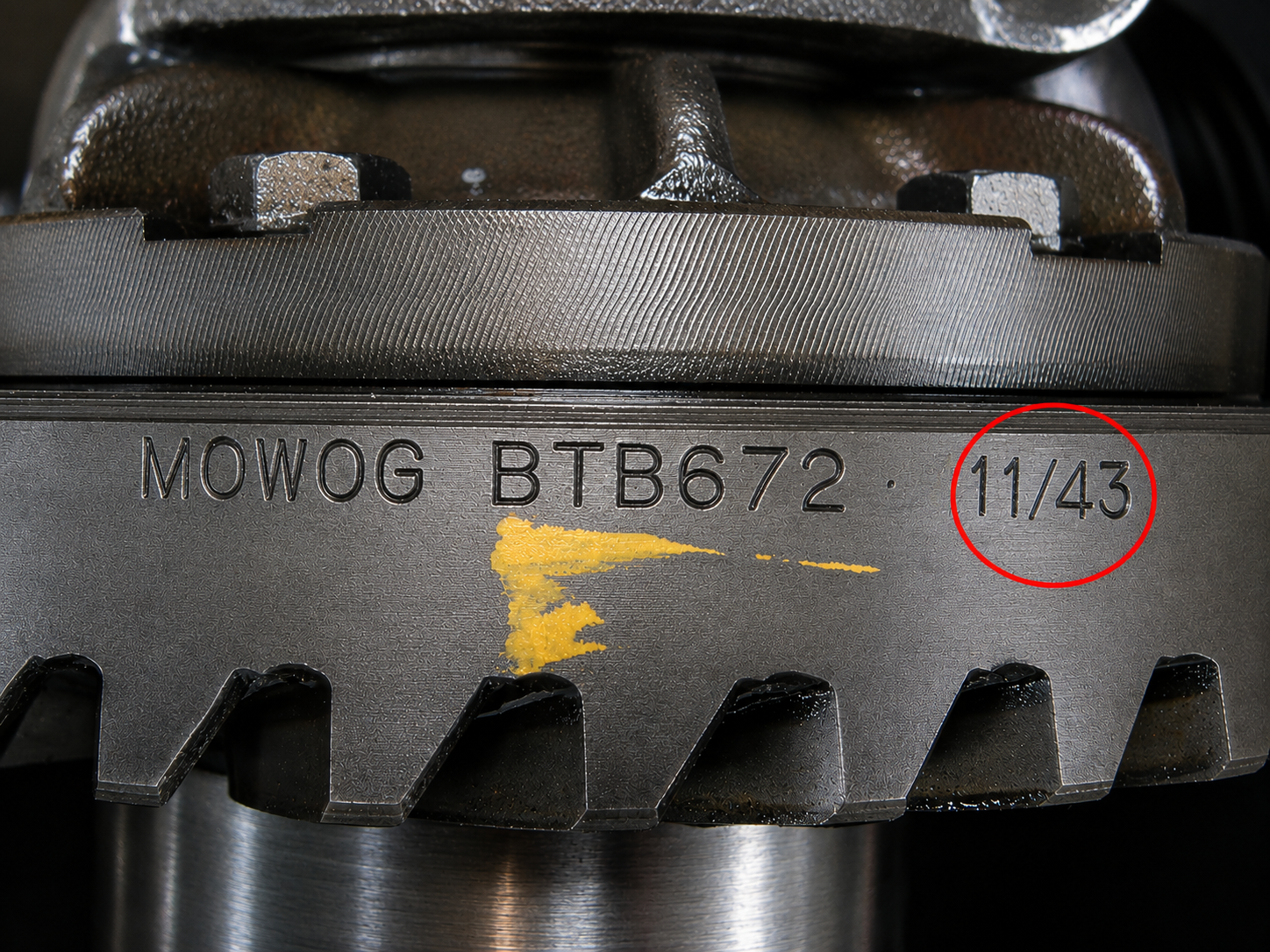 Close-up of a metal gear with the text "MOWOG BTB672" and "11/43" (circled in red) engraved on its surface, along with a yellow paint mark.
