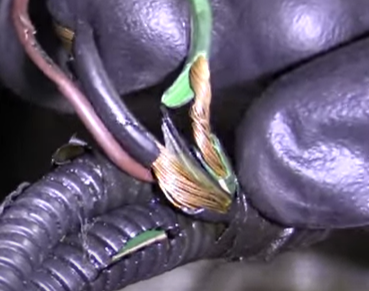 A close-up of a gloved hand holding damaged electrical wires with exposed copper strands and cracked green and black insulation.