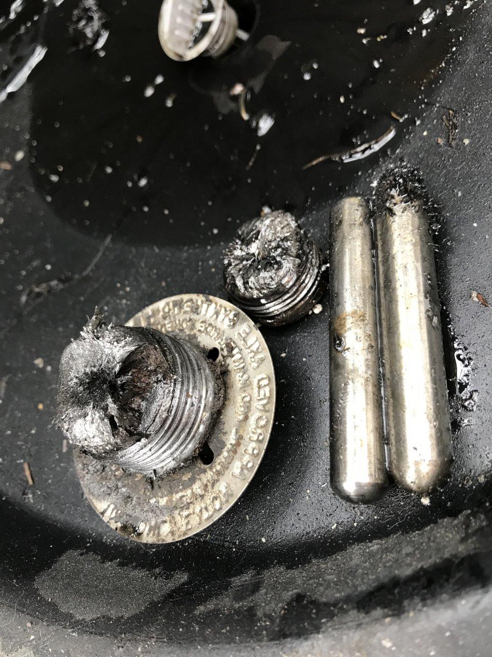 Close-up of burnt and melted metal components, including a threaded cap, a round plate with engraved text, and two cylindrical rods, all resting on a dark surface.