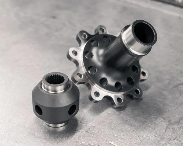 Two precision-machined metal mechanical parts with smooth, polished surfaces are placed on a flat, metallic surface. One part is cylindrical with multiple holes; the other is spherical with internal gear teeth.
