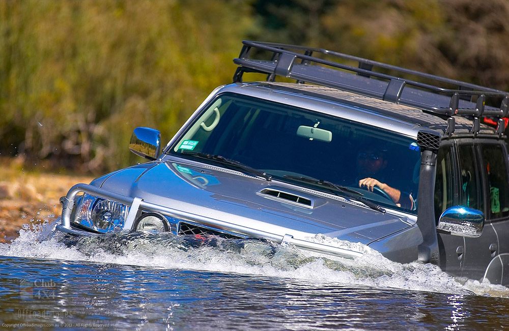A silver SUV with a roof rack drives through deep water, creating splashes around the vehicle. The driver is visible behind the wheel, and trees are blurred in the background.