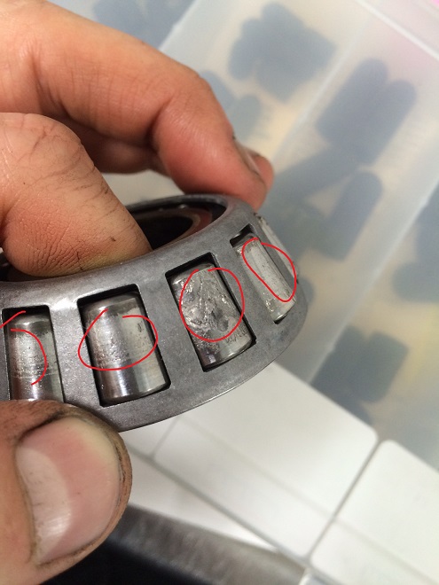 A close-up of a person's hand holding a metal bearing with visible damage and wear marks, highlighted by red circles. The background is out of focus.